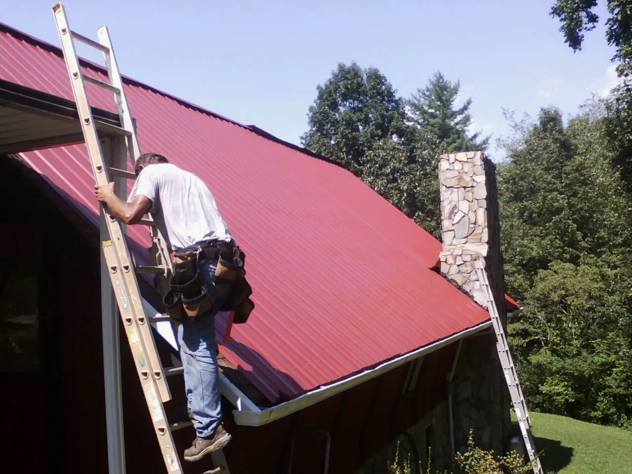 Roofer on a ladder beside a red metal roof and stone chimney — Soffit & Fascia Repair by Roofing Brothers in Beatty